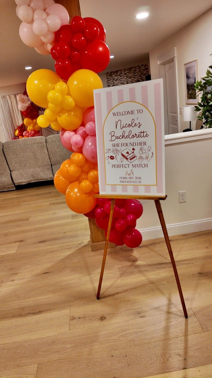 Decorative balloons and a sign on an easel in a room with a couch and decor.