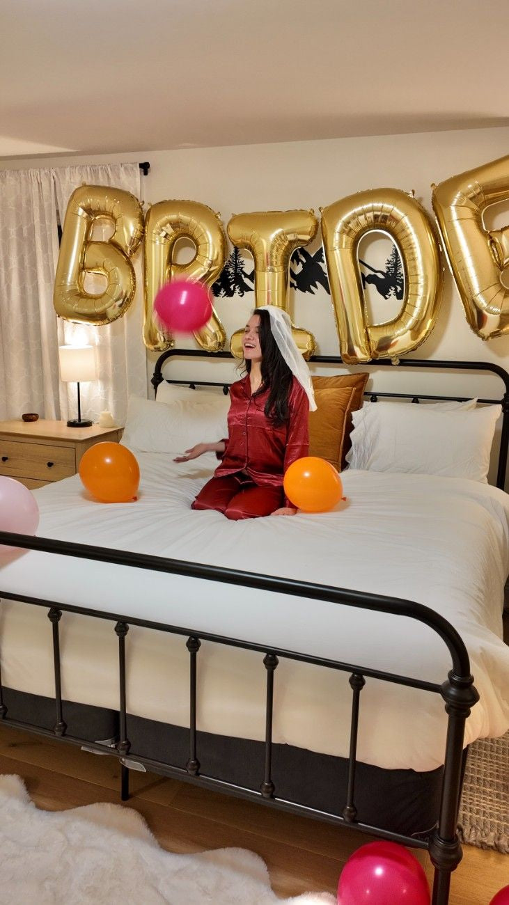 Person in a red dress sitting on a bed with gold 'BRIDE' balloons and colorful balloons.