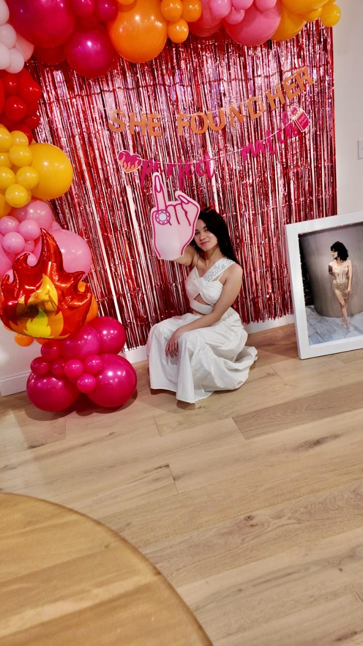 Woman in a white dress sitting in front of a decorative setup with balloons and a photo frame.