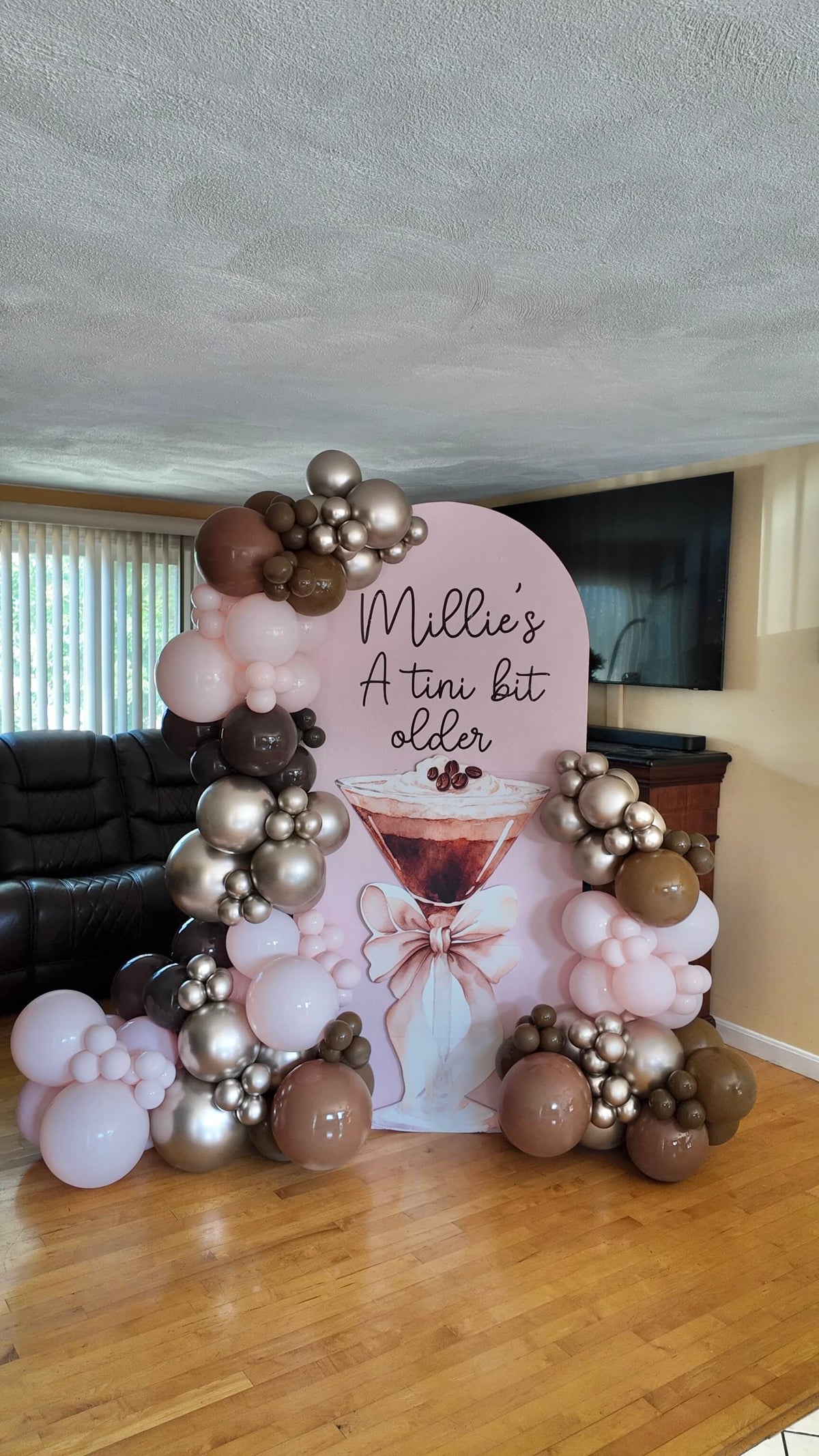 A custom party backdrop stand with a pink arch of balloons in shades of pink, brown, and metallic gold. The backdrop features a large pink arch with the text 'Millie's A tin bit older' and an illustration of a pink cocktail. rhode island balloon decor