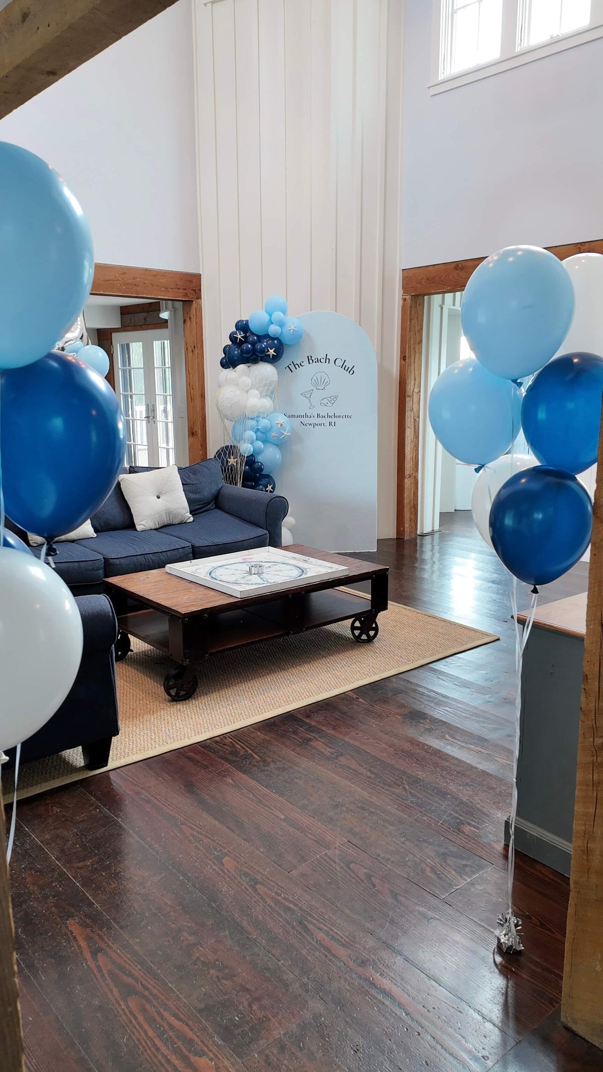 A styled event space with blue and white balloon arrangements framing a living room setup with a dark blue sofa, wooden coffee table, and a white backdrop board.