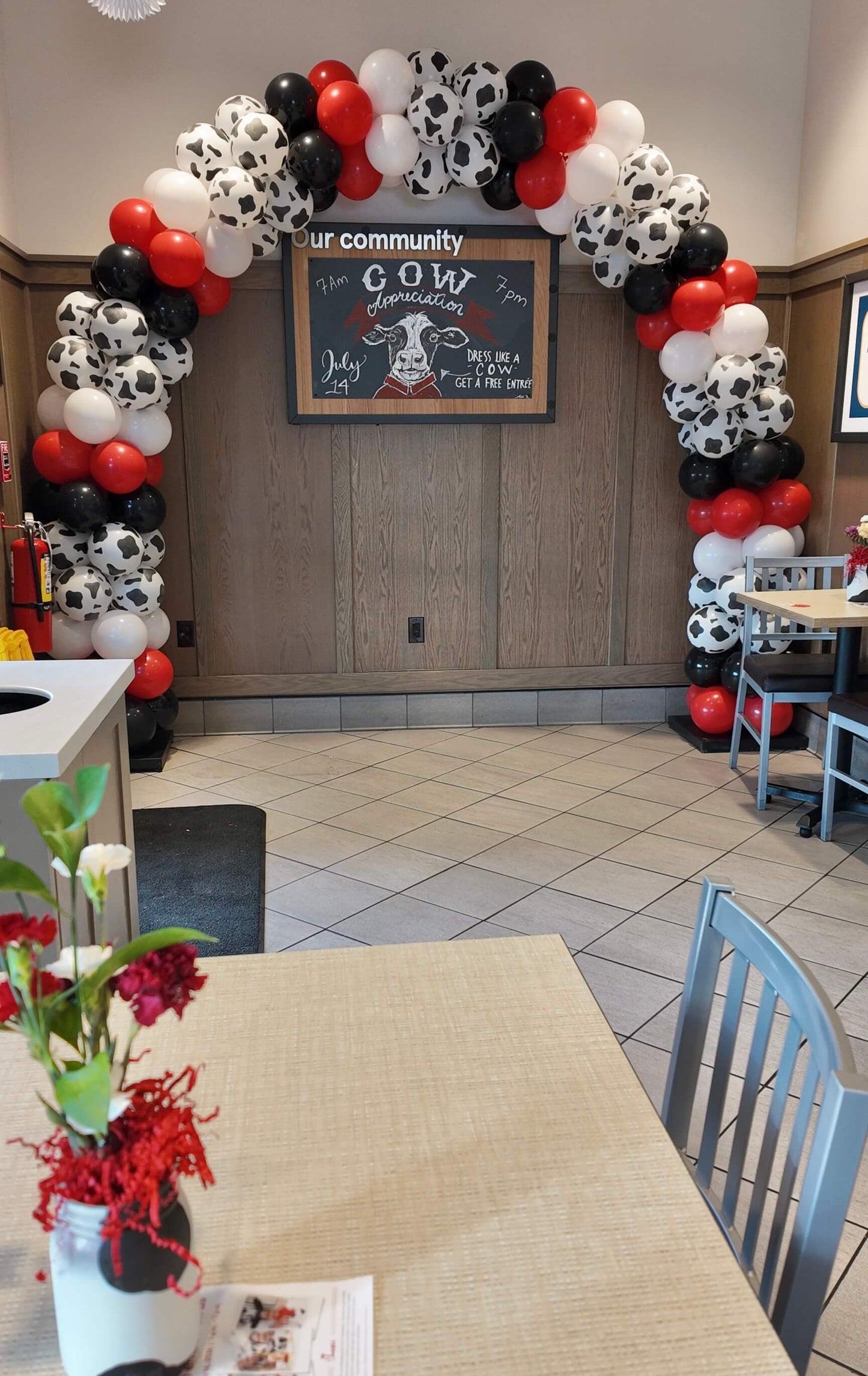 A black, white, and red balloon arch decorated with cow print balloons frames a 'GOW' sign in a restaurant or event space with tiled floors and tables.chickfila balloon arch