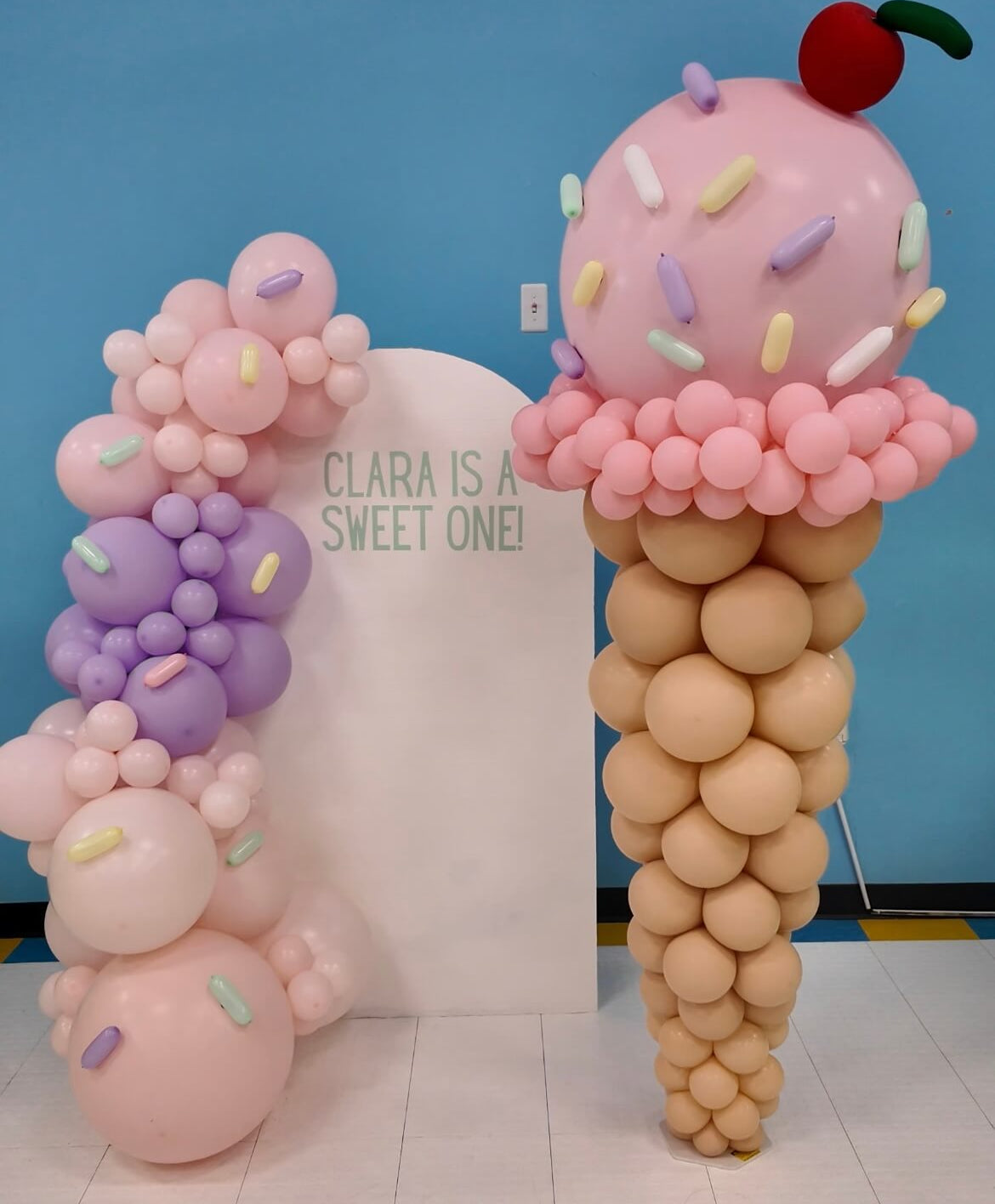 Balloon sculpture of an ice cream cone with sprinkles and a cherry against a blue wall.