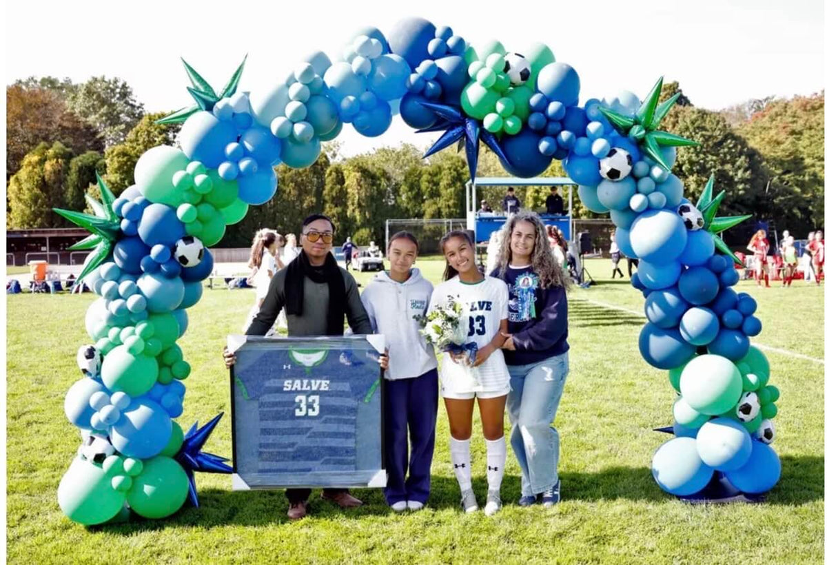 Four people standing on a grassy field under a large blue and green balloon arch decorated with soccer balls and star balloons. One person holds a blue plaque that reads 'SALVE 33'.